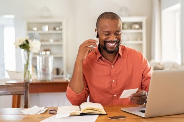 Man sitting and on a cell phone call smiling while looking at a piece of paper.